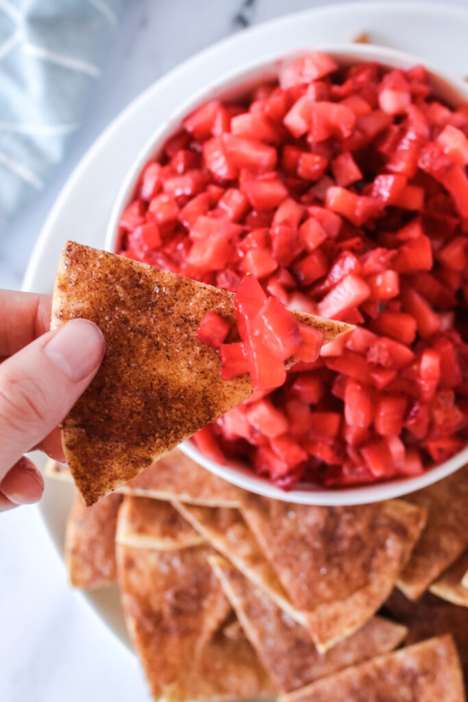 Image taken from the top looking down. Cinnamon sugar tortilla chips with a hand holding one with chopped strawberries on chip.